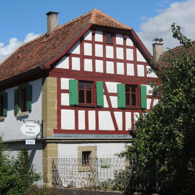 Fachwerkhaus mit braunem/rotem Fachwerk und grünen Fenstern, Fensterläden. Das Haus wurde kürzlich renoviert. Das Haus ist weiß. Am Haus hängt ein Schild mit der Aufschrift "Herberg im Hofhaus".