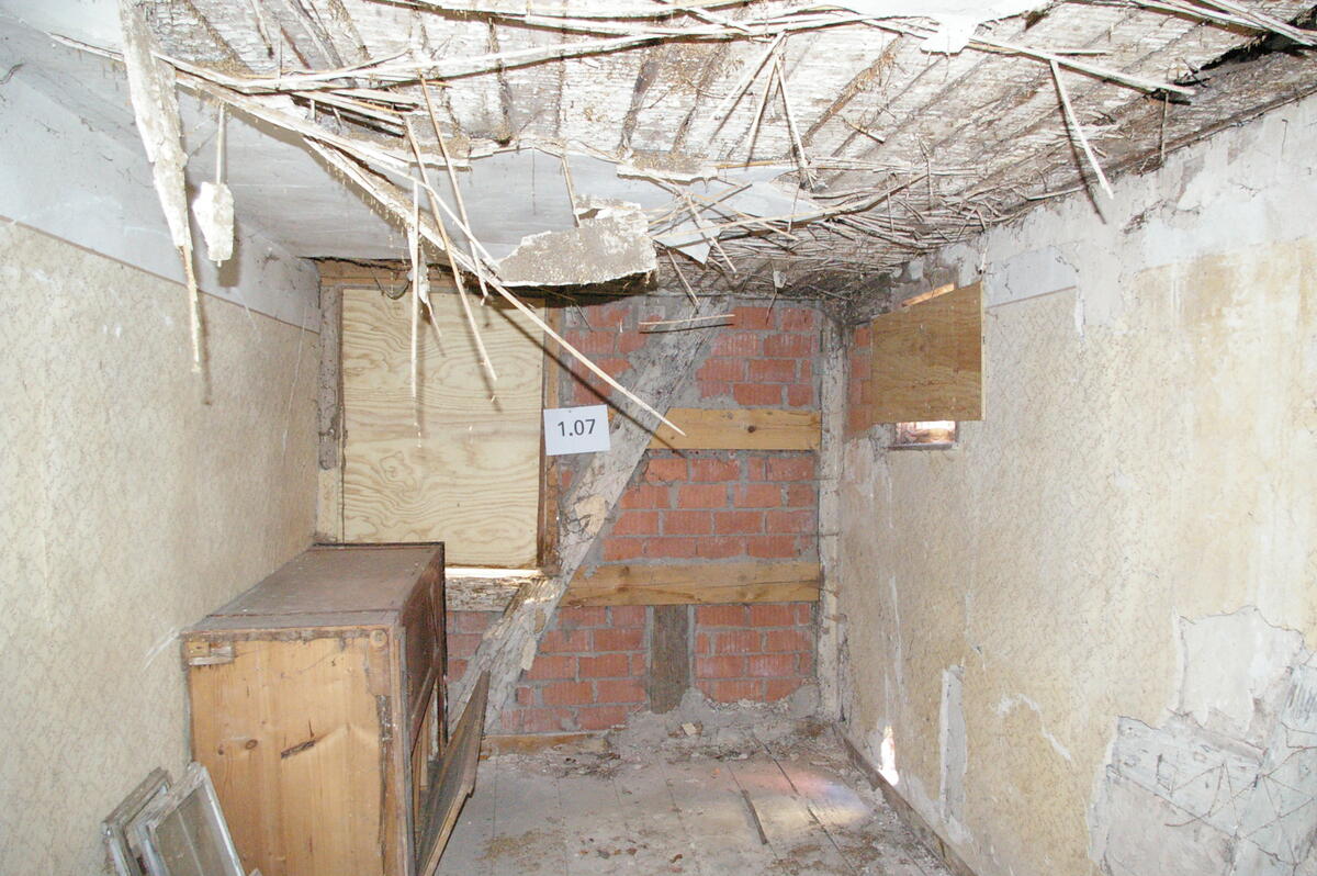 Bild vergrößern: Altes Zimmer mit einer zur Hälfte herausgebrochenen Decke. Eine Wachswand mit Backsteinen zwischen dem Fachwerk. Die beiden anderen Wände sind verputzt, aber der Putz bröckelt ab, der Fußboden ist aus Holz. In der Stube stehen eine alte Kommode und zwei Fensterläden.