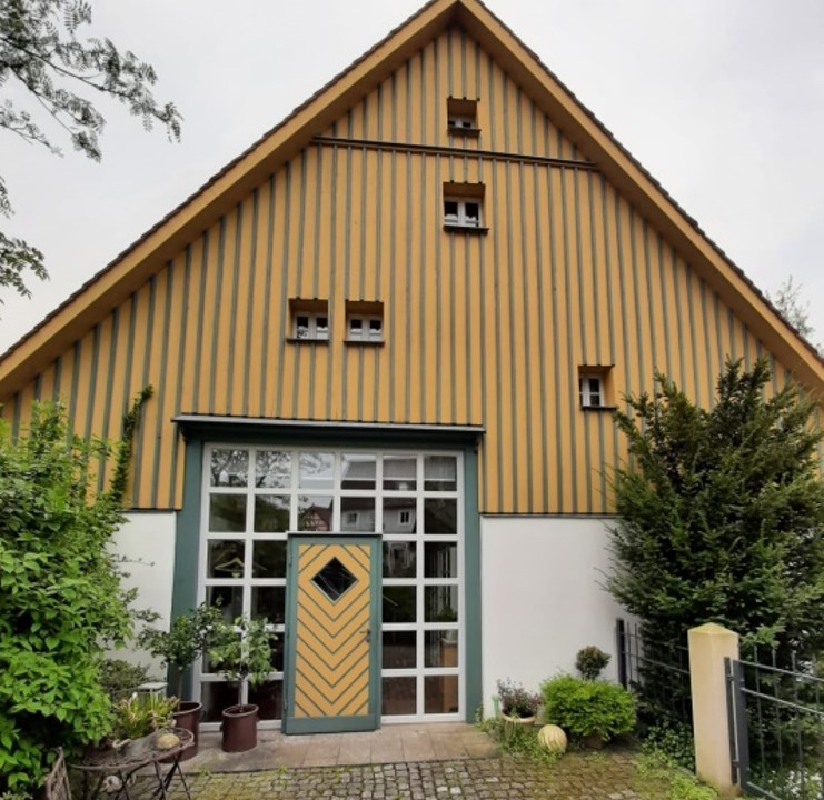 Bild vergrößern: Gelb mit grünen Streifen Holzhaus mit großer Fenstertür. Die Tür ist in der Mitte gelb und grün gestreift, die Fenster um die Tür herum sind weiß. Vor dem Haus sind auf beiden Seiten grüne Büsche.