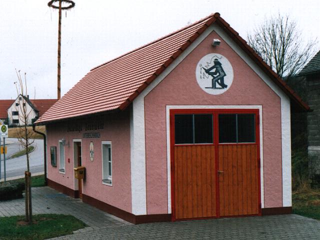 Bild vergrößern: Rosanes Feuerwehrhaus. Das Feuerwehrhaus hat ein großes Garagentor aus Holz mit rotem Rahmen, über dem Garagentor ist ein Bild von einem Feuerwehrmann gemalt.
