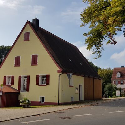 Gelbes Haus mit weißen Fenstern und roten Fensterläden. 
Das Haus hat ein großes Garagentor aus Holz. Das Haus steht direkt an der Straße.