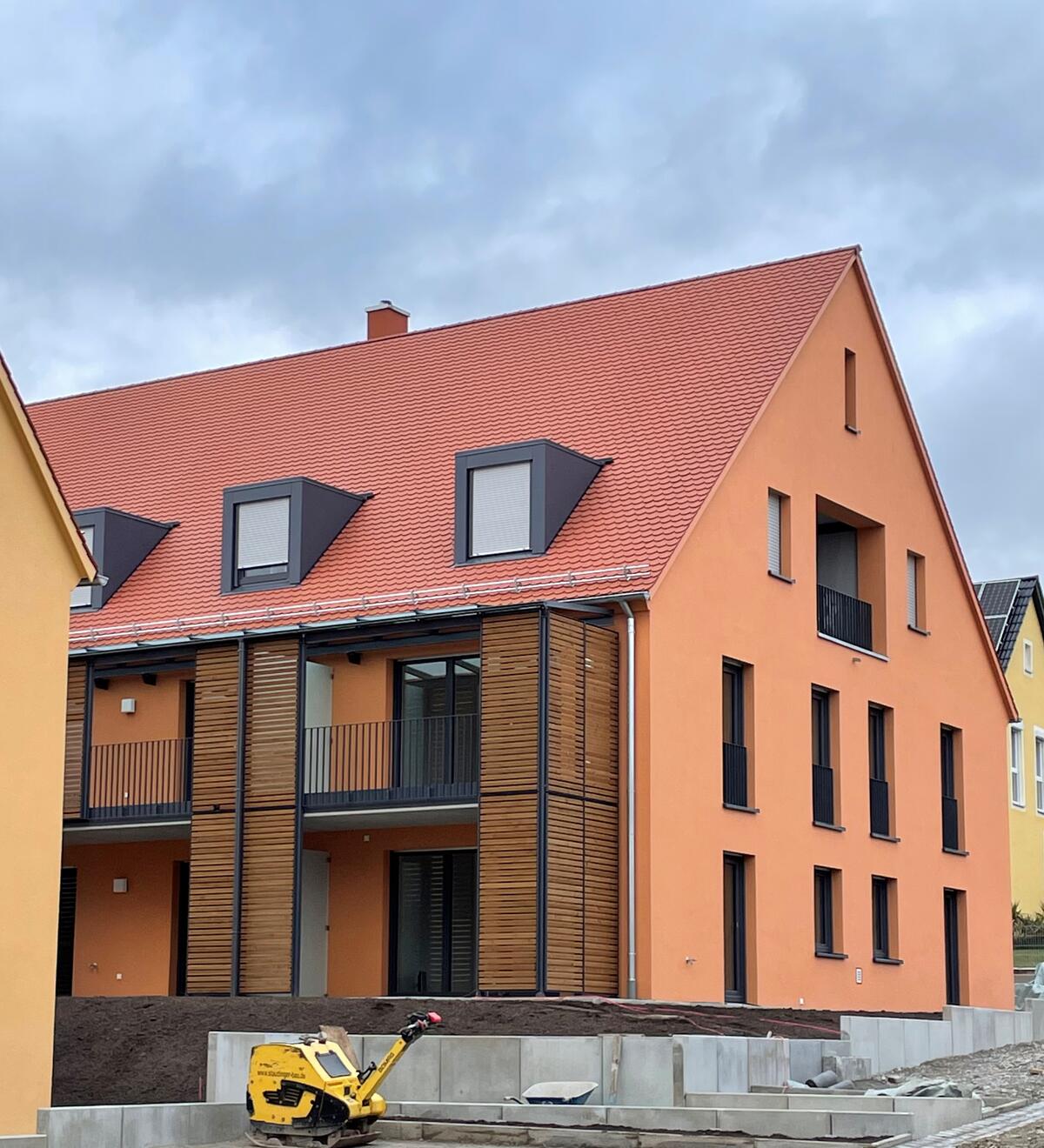 Bild vergrößern: Orangefarbenes Haus mit braunen Fenstern und Türen. Das Haus hat einen schwarzen Balkon mit einer dekorativen Holzwand.