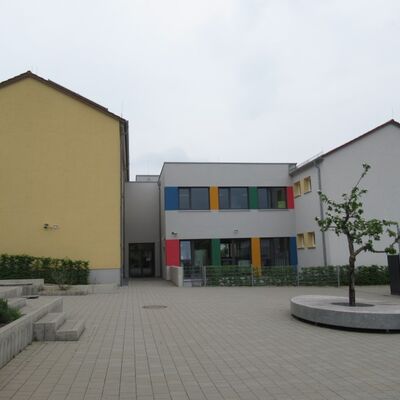 Schule mit zwei Gebäuden. Das eine Gebäude ist grau mit orangen, blauen und roten Akzenten.