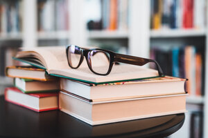 Brille auf einem Bücherstapel. Bibliothek im Hintergrund.