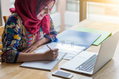 Bild vergrößern: Muslimische Frau, die mit Computer arbeitet und Notizbuch schreibt.